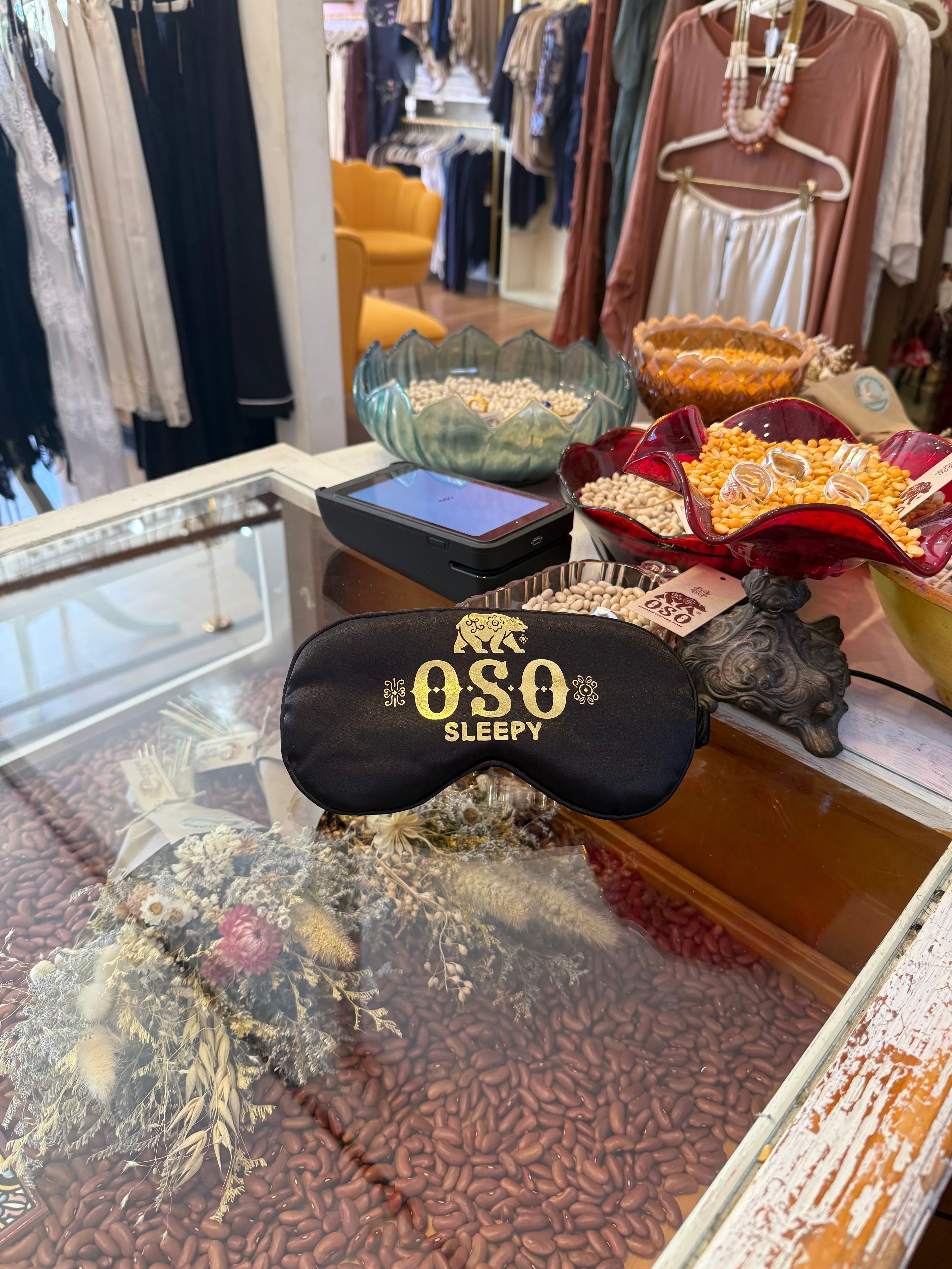 A black sleep mask with "OSO SLEEPY" printed on it rests on a glass display table surrounded by decorative bowls and coffee beans.
