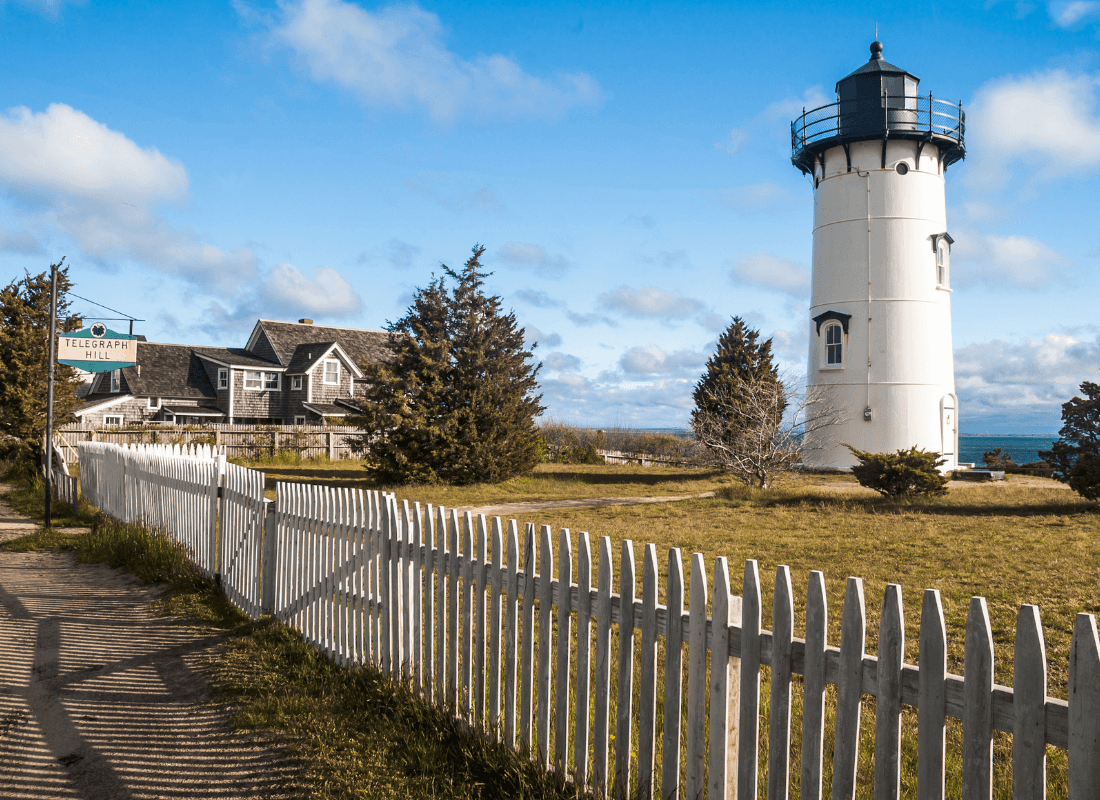 A white lighthouse stands beside a quaint house and a picket fence under a blue sky.