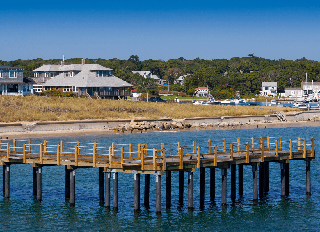 A wooden pier extends into a calm blue sea, with coastal houses and greenery in the background.